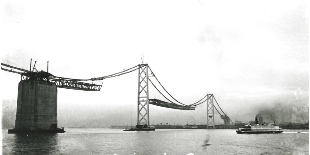 A vintage black-and-white photo from the 1930s shows the Bay Bridge under construction.