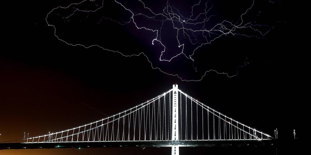 The Bay Bridge is illuminated against the night sky as lightning strikes overhead.