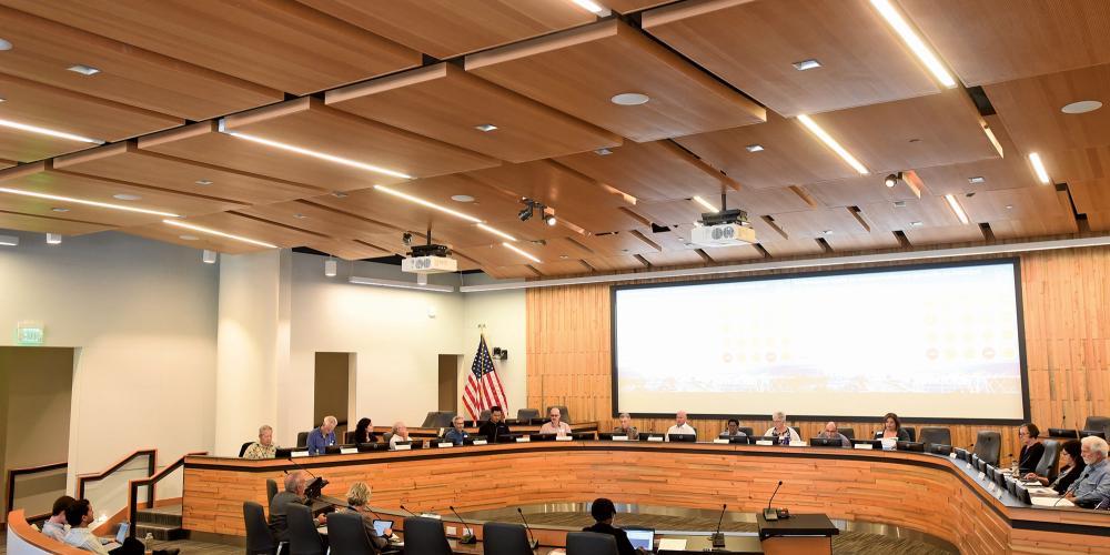 16 people sit at the board seats in the board room of the Bay Area Metro Center in San Francisco. A large white screen and an American flag are behind them,