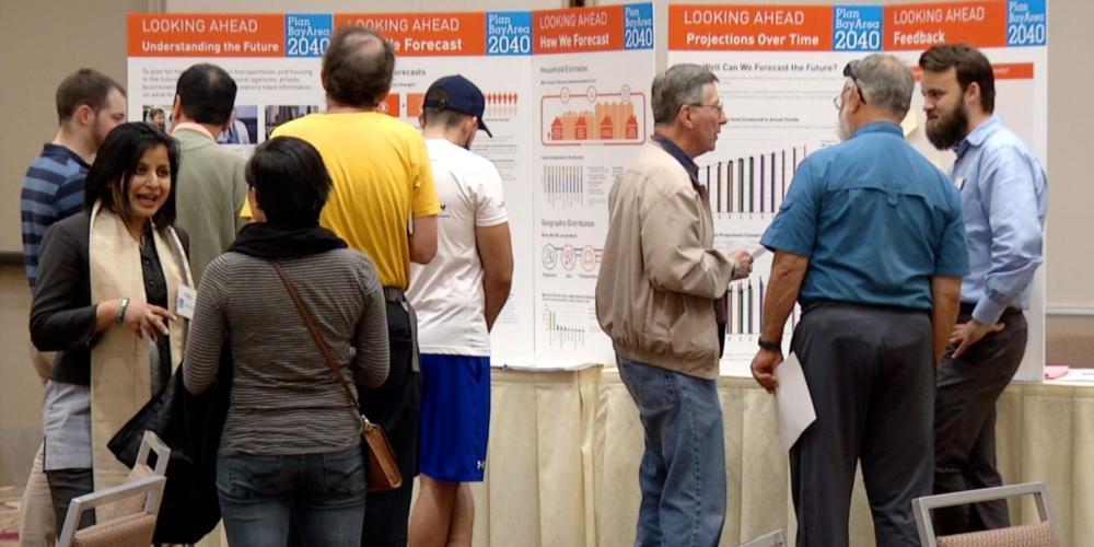 MTC/ABAG staff and members of the public discuss the projections for population and housing for Plan Bay Area 2040 at an open house event. Individuals stand in groups while display boards are on the table in the background.
