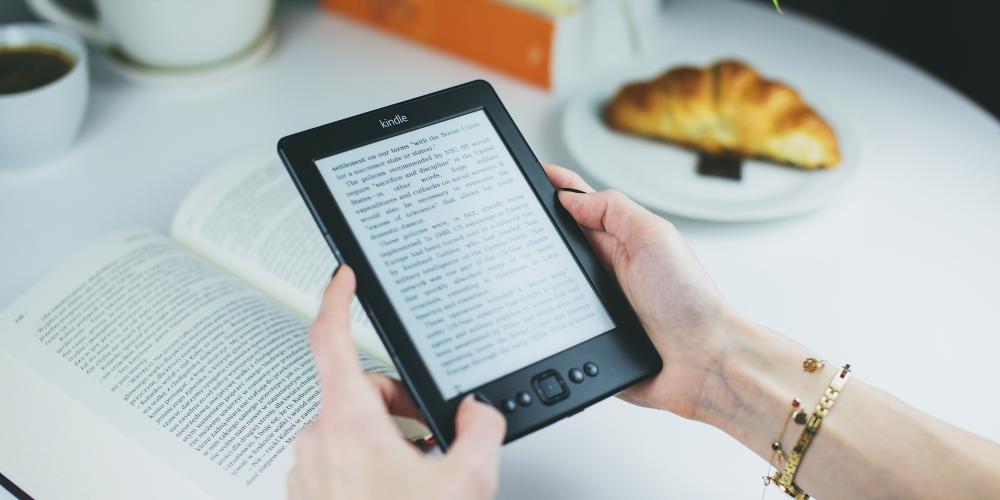 Hands hold a tablet with text on it; a large book is below the tablet. A cup of coffee and a croissant are in the background.