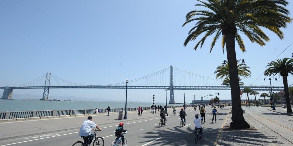 Cyclists and rollerbladers ride along the Embarcadero in San Francisco on a sunny day with the Bay Bridge in the background.  