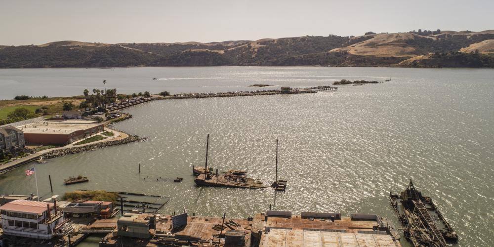 Suisun Bay. The water is a blue-green-brown color; there are some docks and a boat in the foreground, and brown hills covered in dark green trees in the background.