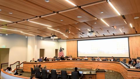 16 people sit at the board seats in the board room of the Bay Area Metro Center in San Francisco. A large white screen and an American flag are behind them,