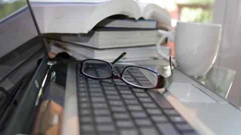 A pair on glasses rests on a laptop keyboard. Several books and a coffee cup are shown to the side of the laptop.